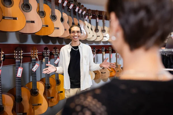 Smiling african american seller pointing with hands near acoustic guitars and blurred customer in music store