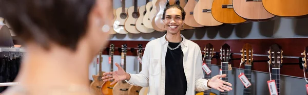 African american seller pointing with hands near acoustic guitars and blurred customer in music store, banner