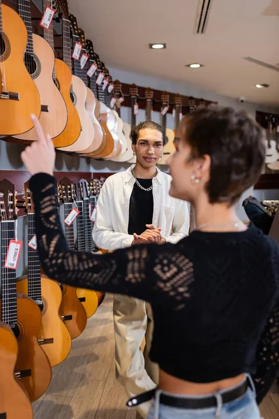 African american seller standing near blurred customer pointing at acoustic guitars in music store
