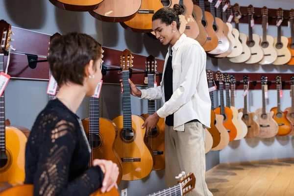 Positive african american seller holding acoustic guitar near blurred customer in instrumental music store