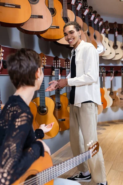 Positive african american seller pointing at acoustic guitar near woman in music shop