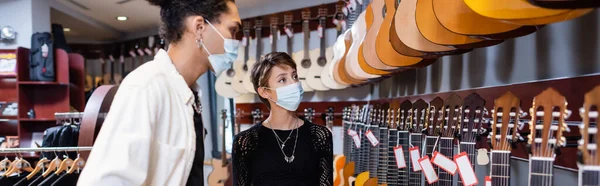 Young woman in medical mask choosing acoustic guitar near african american seller in music store, banner