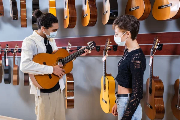 African american seller in medical mask playing acoustic guitar near customer in store