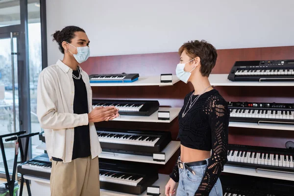 African american seller in medical mask looking at customer near synthesizers in music store