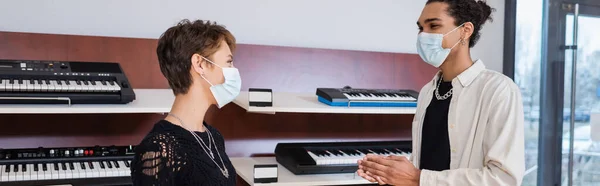 African american seller and customer in medical masks standing near synthesizers in music shop, banner