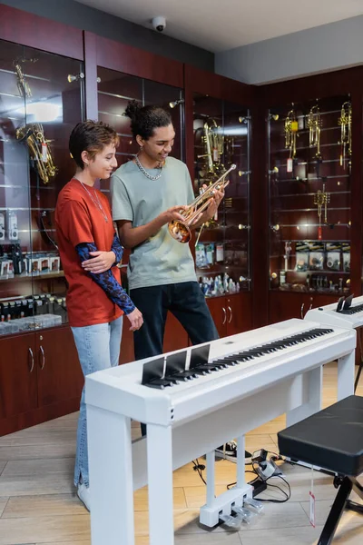 Interracial seller and customer looking at trumpet near piano in musical store