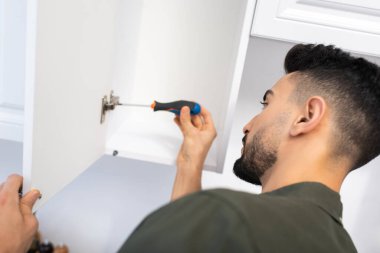 Side view of arabian man fixing hinge in blurred kitchen cabinet at home 