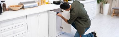 Young muslim man with screwdriver fixing cabinet under worktop in kitchen, banner 