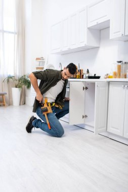 Arabian man looking at tool belt near cabinet in kitchen 