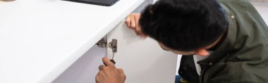 Blurred man with screwdriver fixing door of cabinet in kitchen, banner 