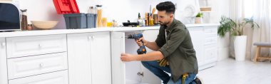 Muslim man with electric screwdriver fixing door of kitchen cabinet, banner 