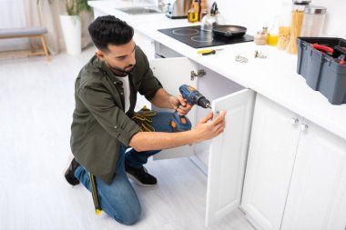Arabian man with electric screwdriver fixing cabinet under worktop in kitchen 