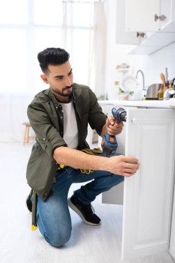 Muslim man with electric screwdriver fixing door of kitchen cabinet 