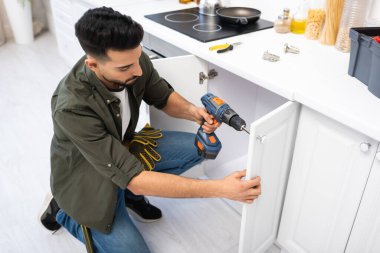 Overhead view of arabian man with electric screwdriver fixing door of cabinet near tools in kitchen 