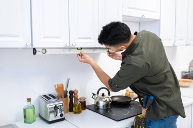 Muslim man checking kitchen cupboard with spirit level near worktop 