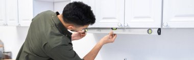 Arabian man measuring kitchen cupboard with spirit level at home, banner 