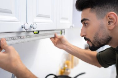 Side view of arabian man looking at spirit level near cupboard at home 