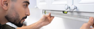 Side view of arabian man measuring kitchen cupboard with spirit level, banner 