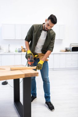 Arabian craftsman in gloves using jigsaw machine on wooden board at home 