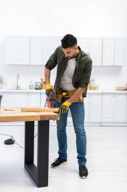 Young muslim craftsman holding jigsaw machine near wooden plank in kitchen 