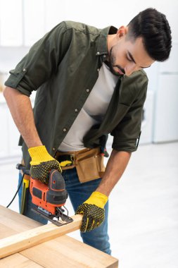 Arabian man with tool belt using jigsaw machine on wooden plank at home 