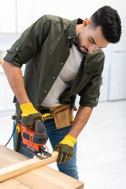 Arabian man with tool belt using jigsaw machine on wooden plank at home  clipart