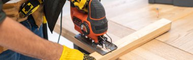 Cropped view of man in gloves using jigsaw machine on wooden plank at home, banner  clipart