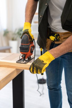 Partial view of man in gloves and tool belt holding jigsaw machine near wooden plank at home 