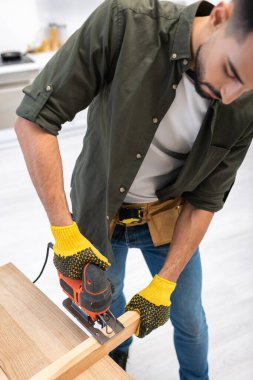 High angle view of young muslim carpenter holding jigsaw machine near wooden board at home 