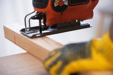 Close up view of blurred man in glove using jigsaw machine on wooden plank 
