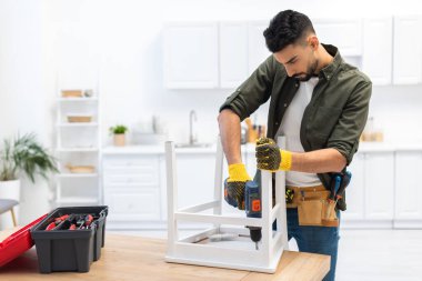 Muslim man in gloves fixing chair with electric drill near toolbox on table at home 