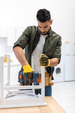 Arabian man in gloves fixing chair with electric drill at home 