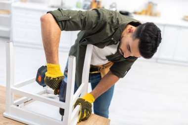 Muslim man in gloves using electric screwdriver while fixing chair in kitchen 
