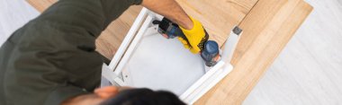 Overhead view of young man repairing chair with electric screwdriver at home, banner  