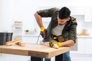 Young muslim man in gloves sanding wooden plank with sander at home 