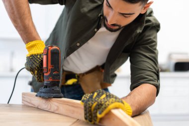 Muslim man in gloves sanding wooden board on table at home 