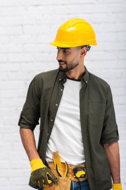 Smiling muslim man in hardhat and gloves touching tool belt at home  clipart