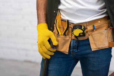 Cropped view of man in glove and tool belt holding hammer at home 