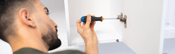 Side view of muslim man with screwdriver fixing kitchen cabinet, banner 