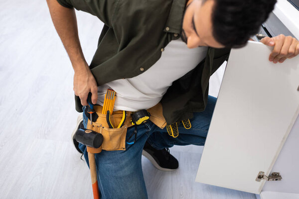 High angle view of arabian man taking screwdriver from tool belt near kitchen cabinet 