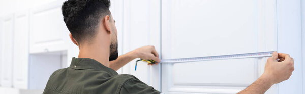 Arabian man with tape measure measuring kitchen cabinet, banner 
