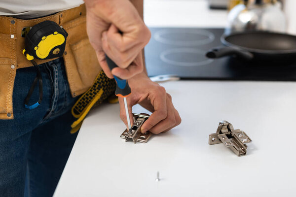 Cropped view of man with tool belt fixing cupboard hinge at home