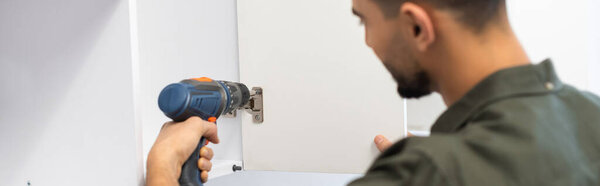 Arabian craftsman with electric screwdriver fixing cabinet in kitchen, banner 