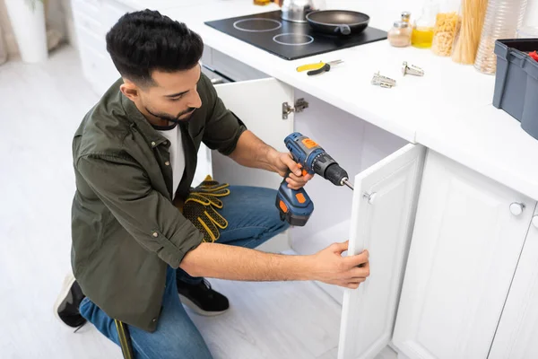 Overhead view of arabian man with electric screwdriver fixing door of cabinet near tools in kitchen