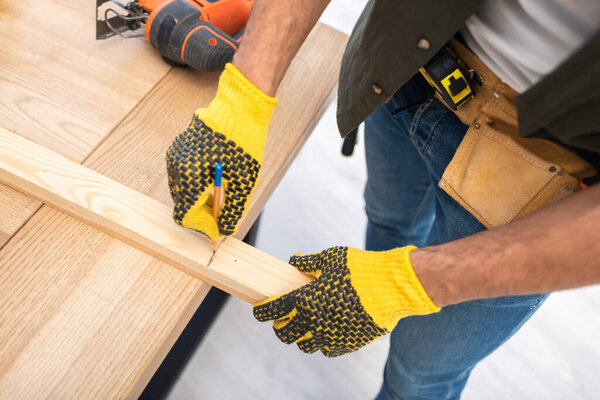 Cropped view of man in gloves and tool belt marking wooden plank at home 