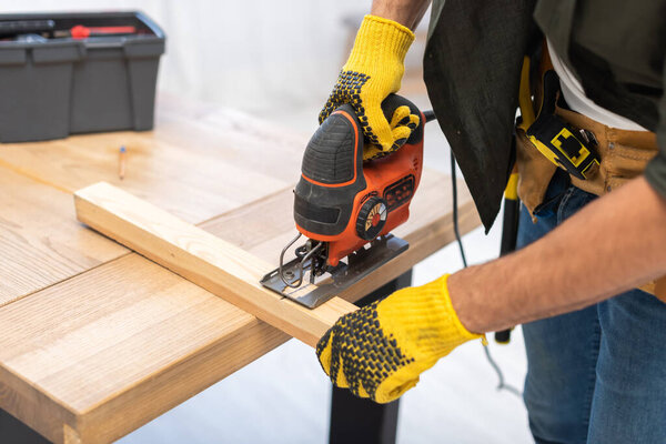 Cropped view of carpenter holding jigsaw machine near wooden plank on table at home 