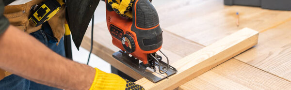 Cropped view of man in gloves using jigsaw machine on wooden plank at home, banner 
