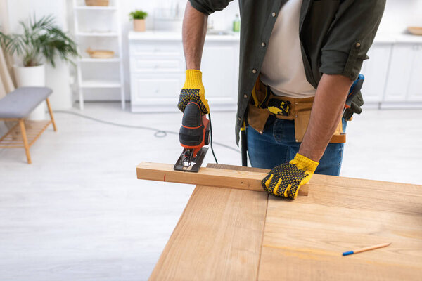 Cropped view of man in gloves holding jigsaw machine while sawing plank at home 