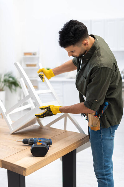 Arabian craftsman in gloves holding chair near electric drill at home 