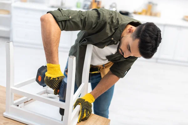 Muslim man in gloves using electric screwdriver while fixing chair in kitchen 